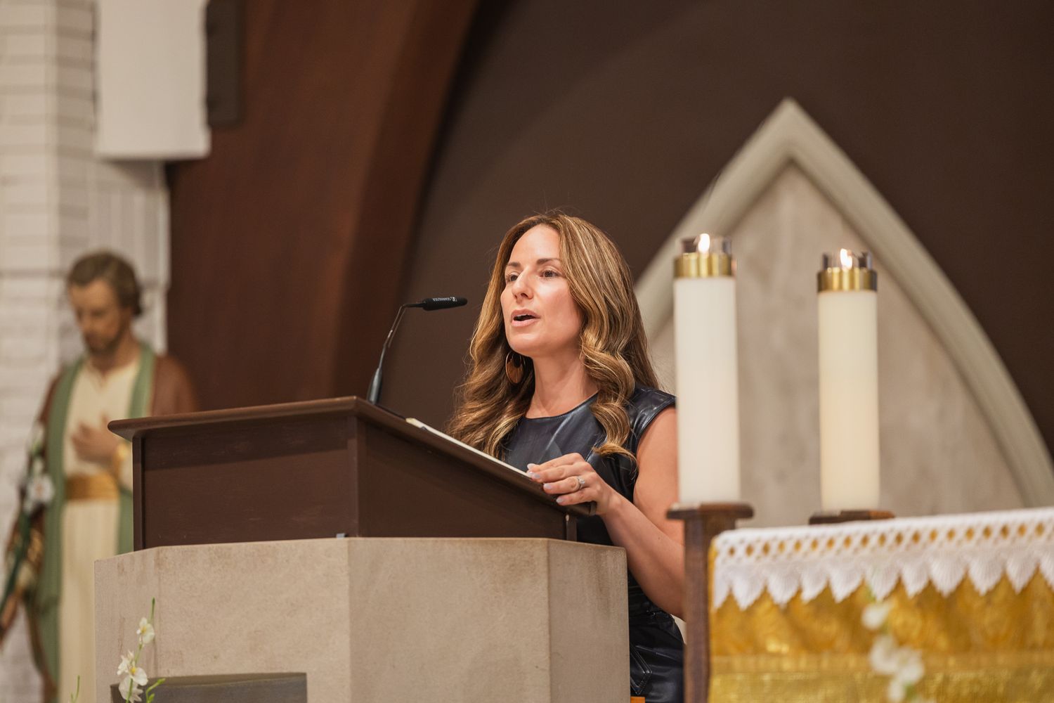 Woman speaking at church podium during wedding ceremony with candles and altar in background.