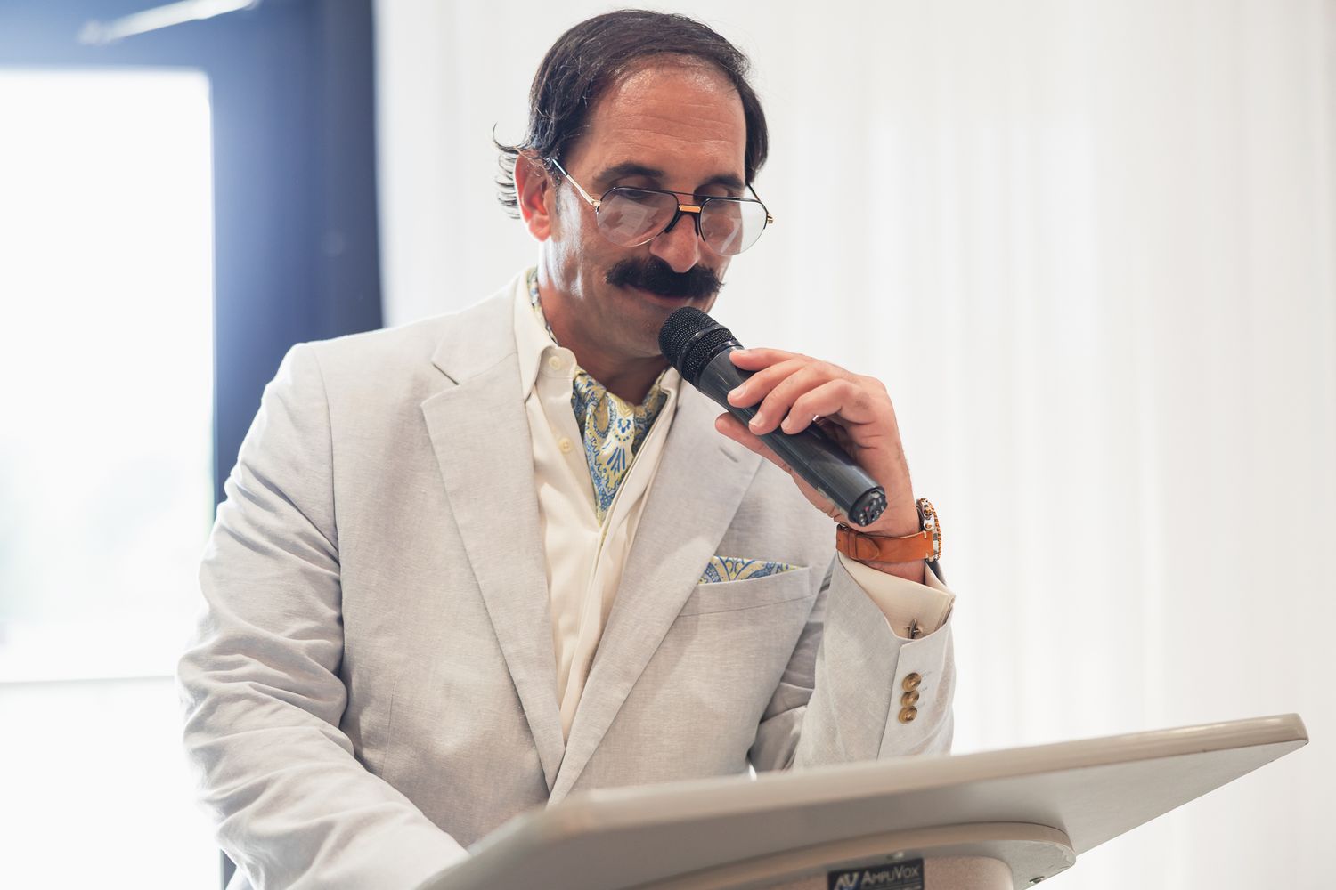 Man in white suit speaking into microphone at podium with distinctive mustache during formal event.