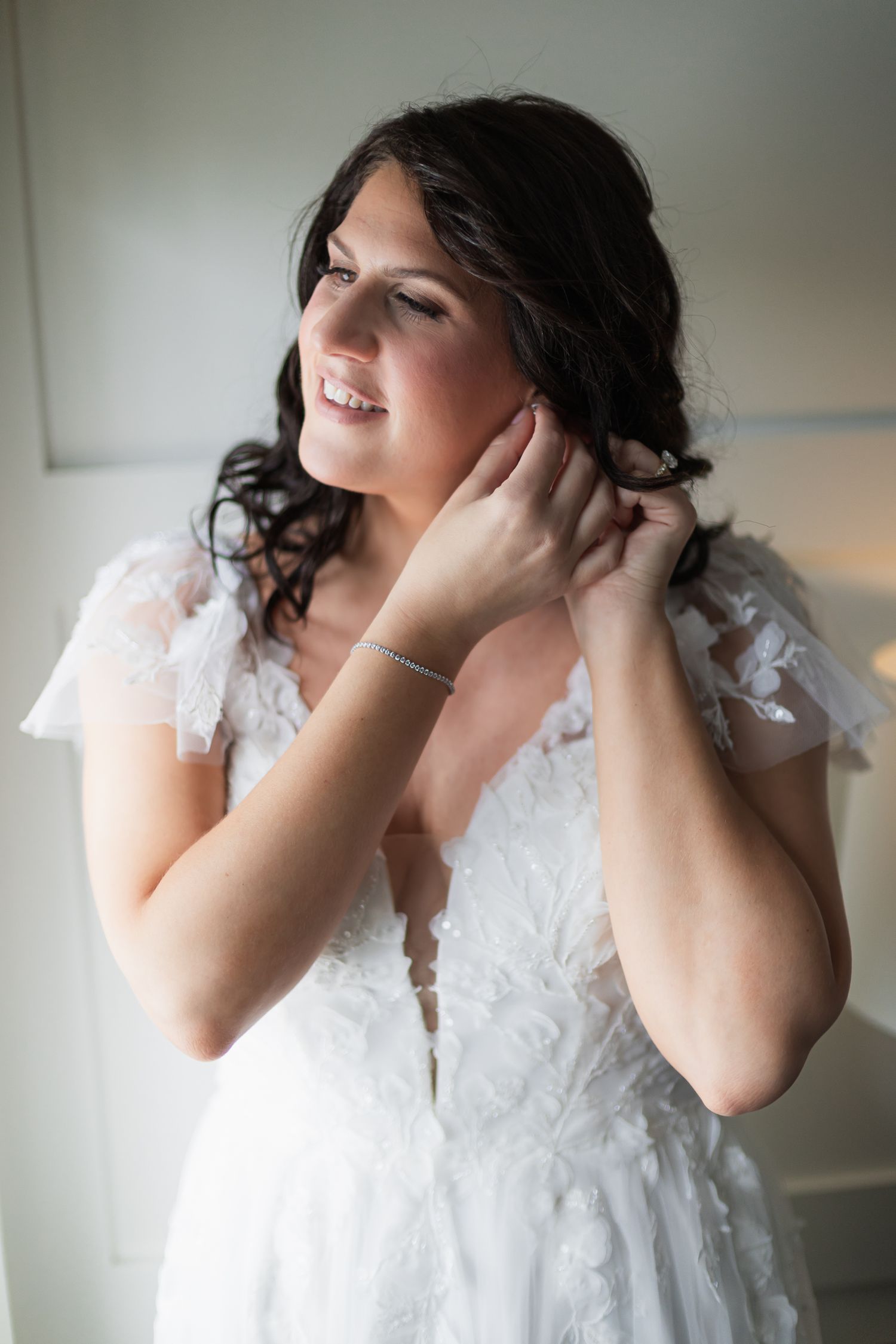 Radiant bride in lace wedding dress smiling while putting on earrings before ceremony.