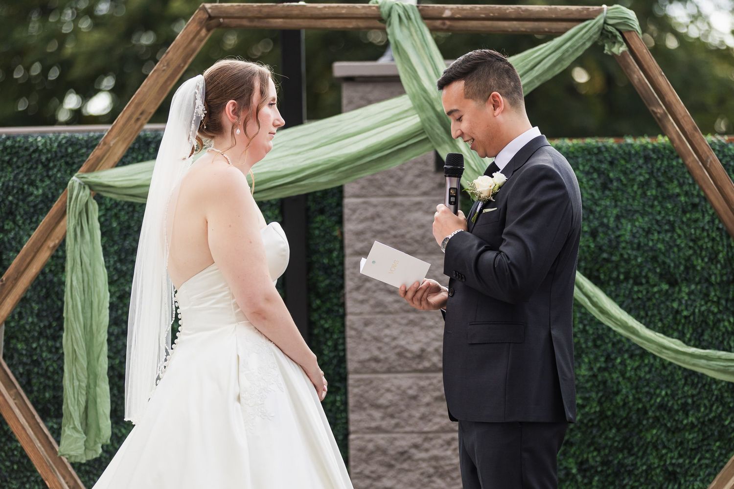 Groom reads vows to bride at outdoor wedding ceremony under geometric arch with sage green fabric.