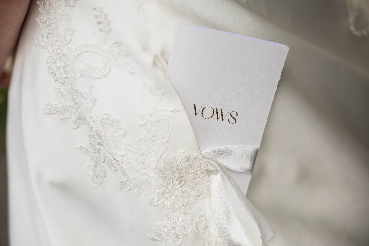 Close-up of bride holding white vows booklet against lace wedding dress detail.