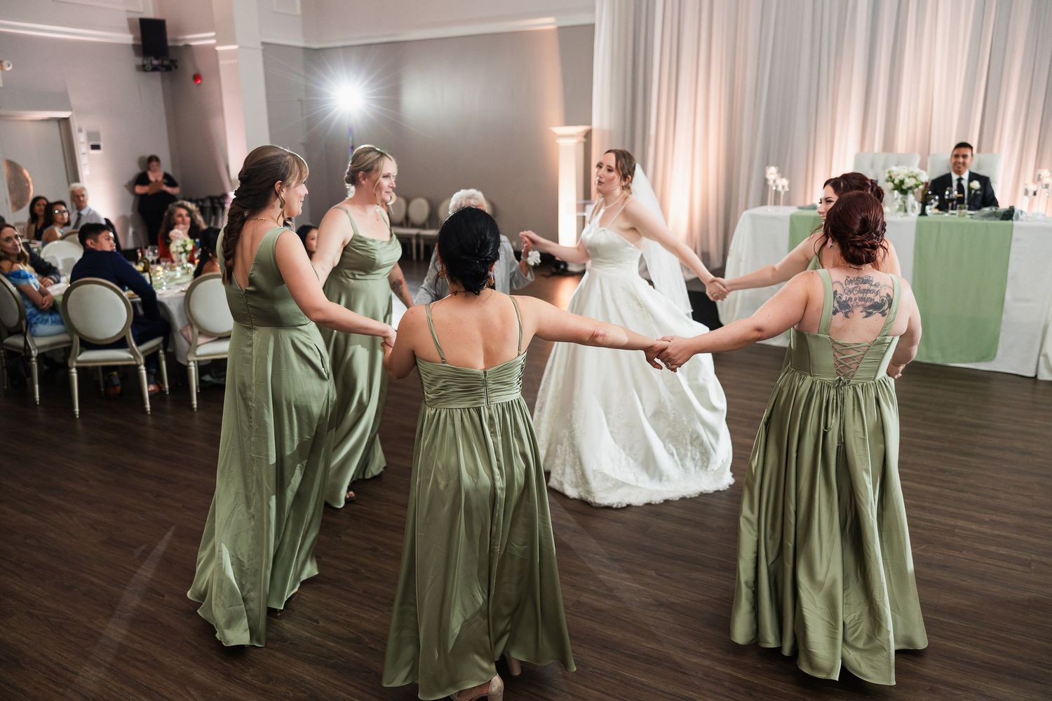 Bride in white gown dances in a circle with bridesmaids in sage green dresses at a wedding reception.