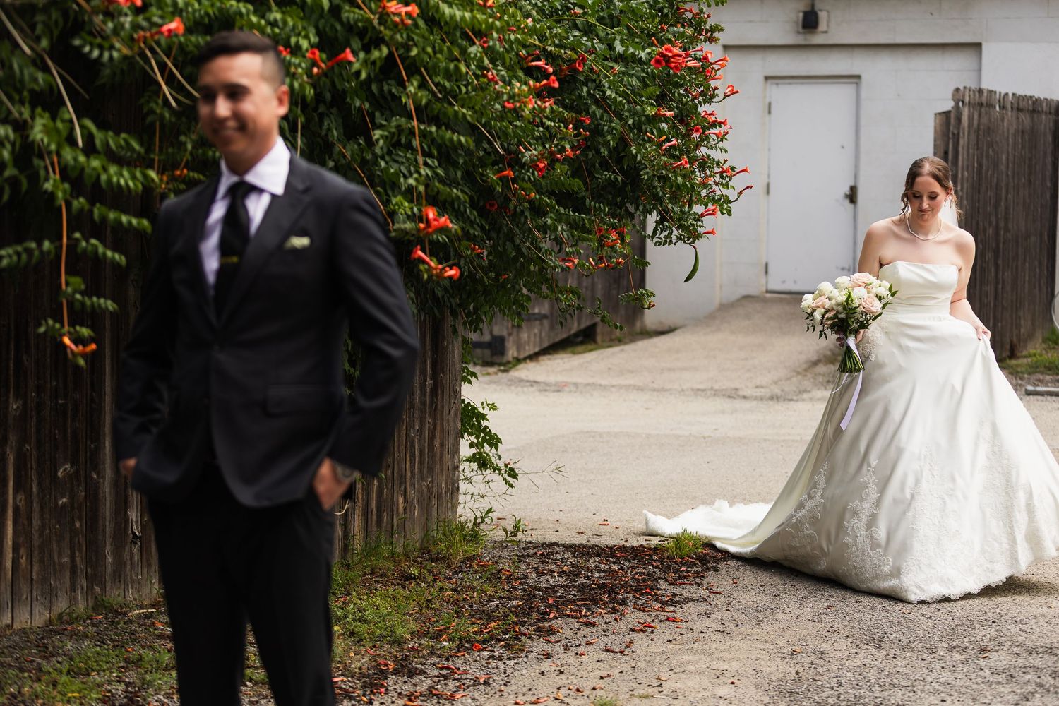 Bride in white gown holds bouquet while groom in black suit stands apart in a romantic outdoor wedding photo.