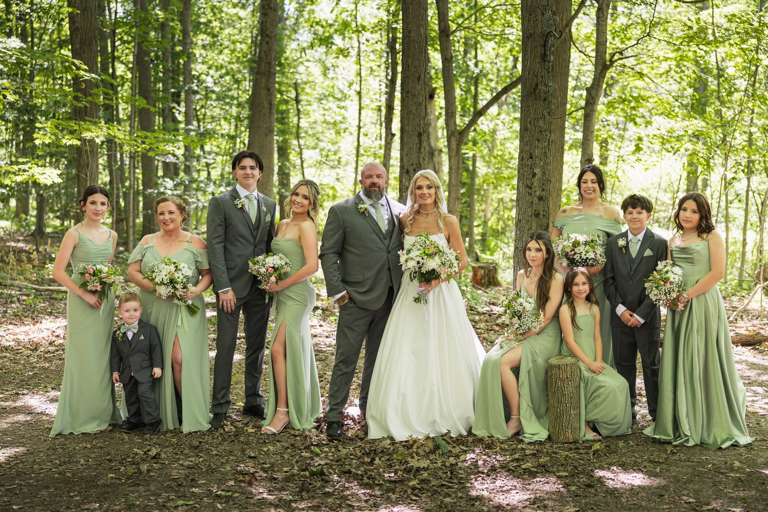 Wedding party in sage green attire poses together in a sunlit forest setting with bride and groom at center.