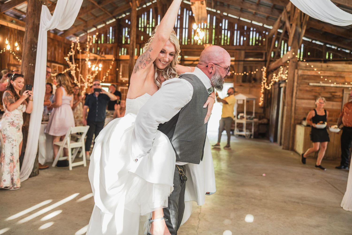 Groom lifting bride in white gown during first dance at rustic barn wedding reception with string lights.