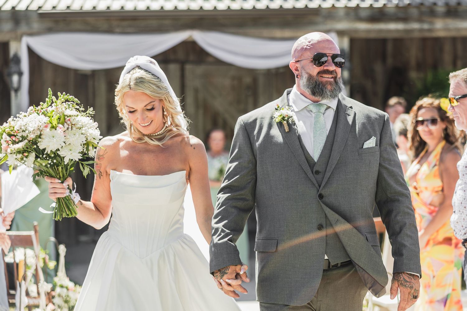 Bride and groom walk hand in hand after outdoor wedding ceremony, bride holding white bouquet in sunny venue.