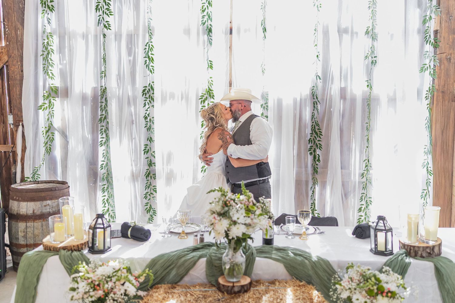 Bride and groom share a kiss at decorated sweetheart table with floral arrangements and white drape backdrop.