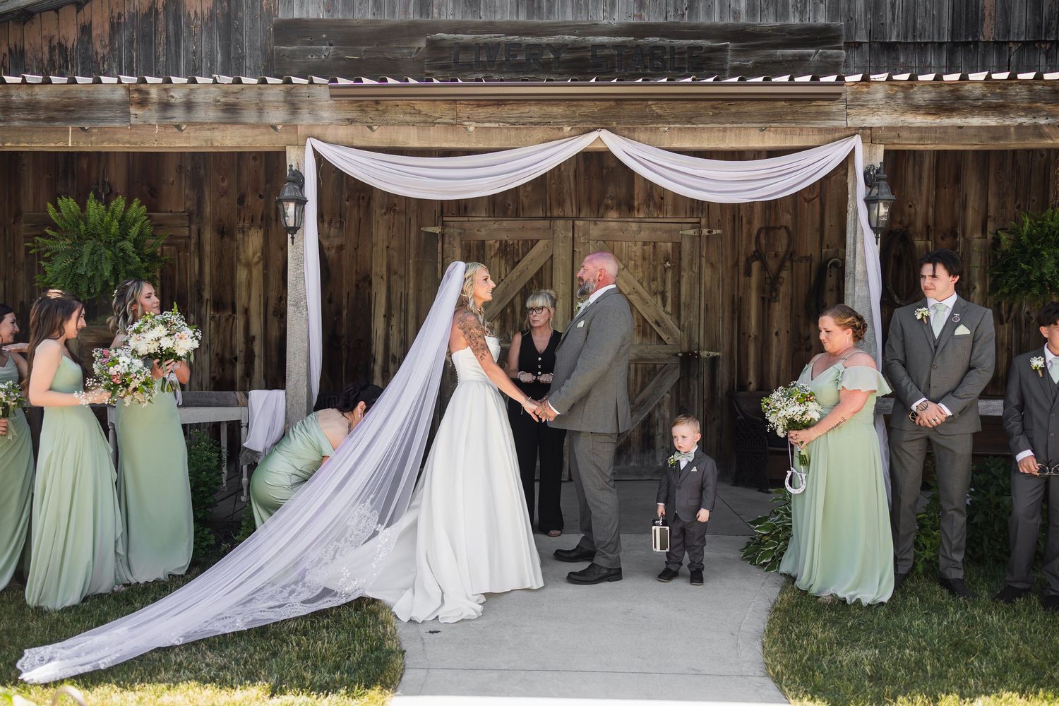 Bride and groom exchange vows under white draped arch at outdoor barn wedding with wedding party.