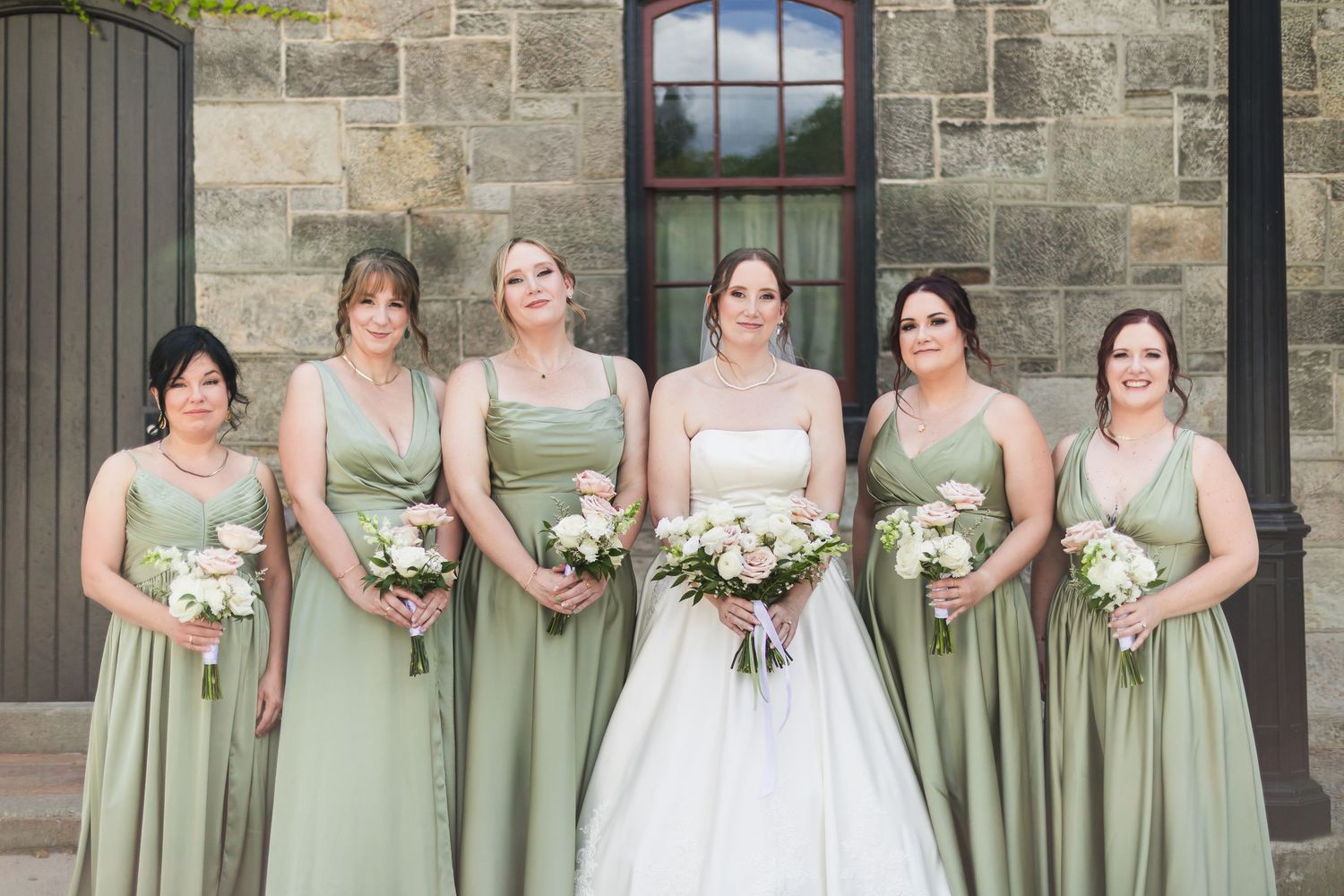 Bride in white gown poses with five bridesmaids in sage green dresses holding bouquets outside stone building.