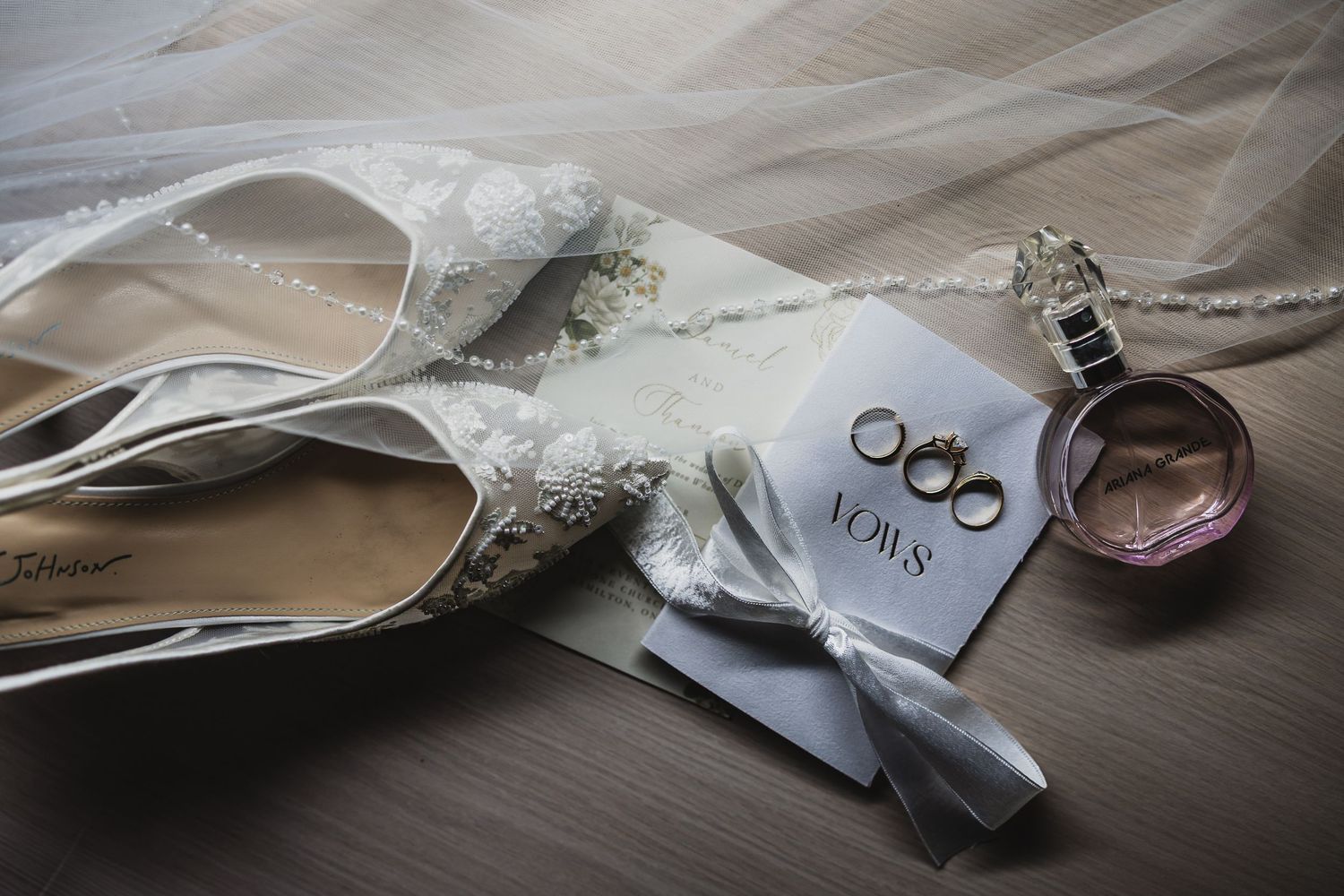 Wedding flat lay featuring bridal heels, gold rings, vows card, and perfume on wood surface.