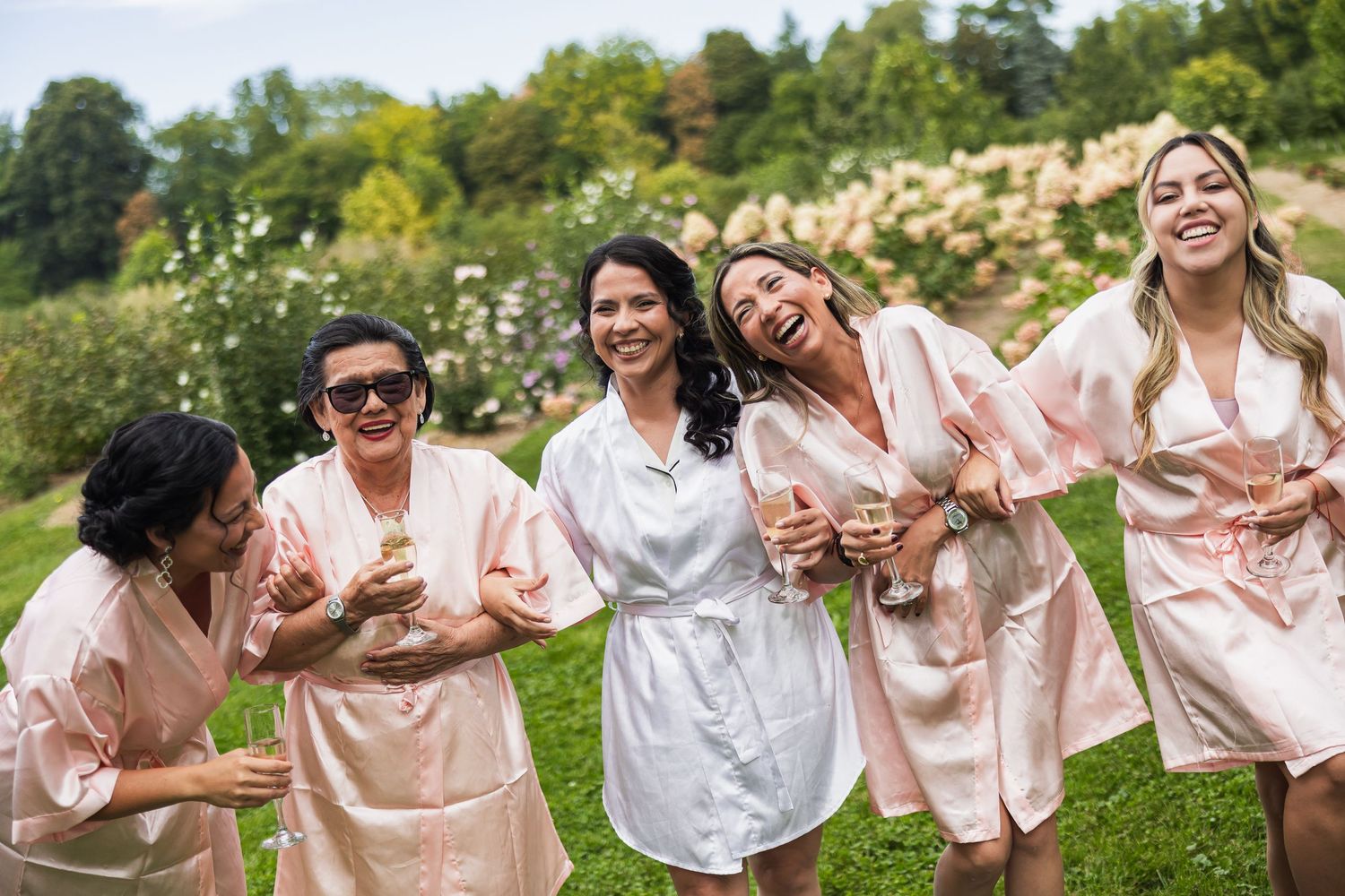 Bride and bridesmaids laughing in matching blush satin robes outdoors before wedding ceremony.