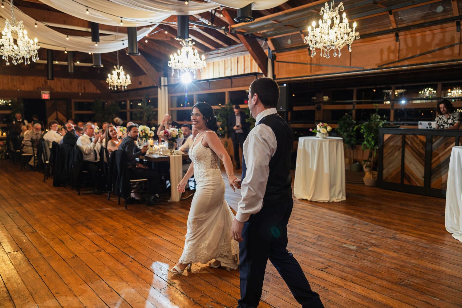 Bride and groom share first dance on wooden floor at elegant rustic wedding reception with chandeliers.