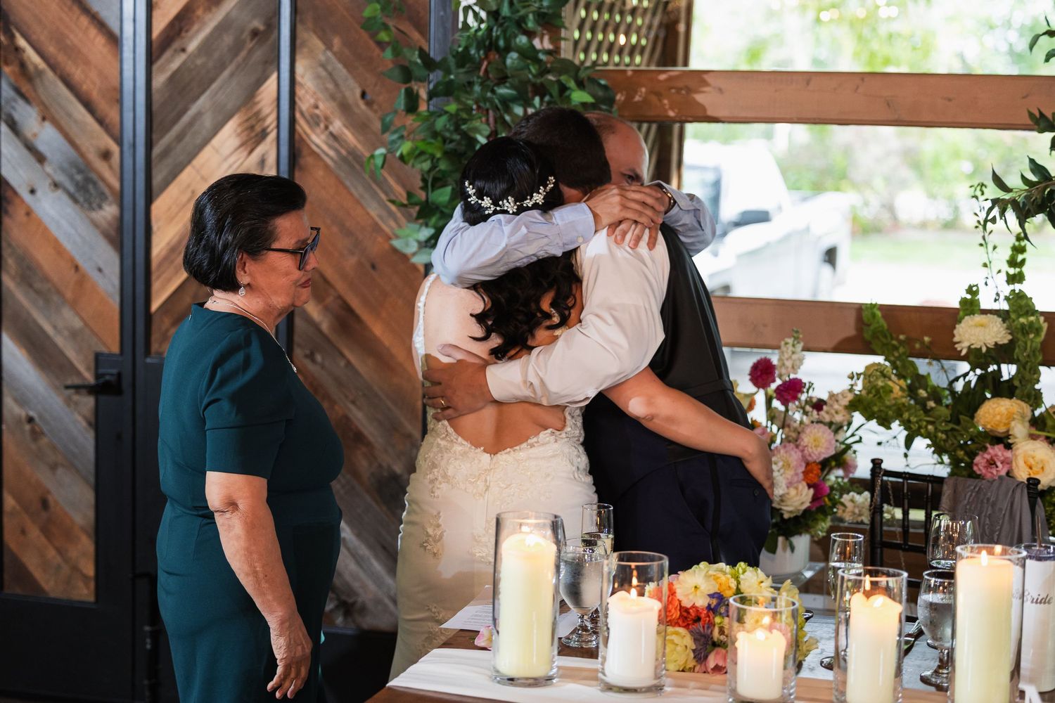 Bride and groom share a heartfelt hug at the reception table while a guest in teal looks on smiling.