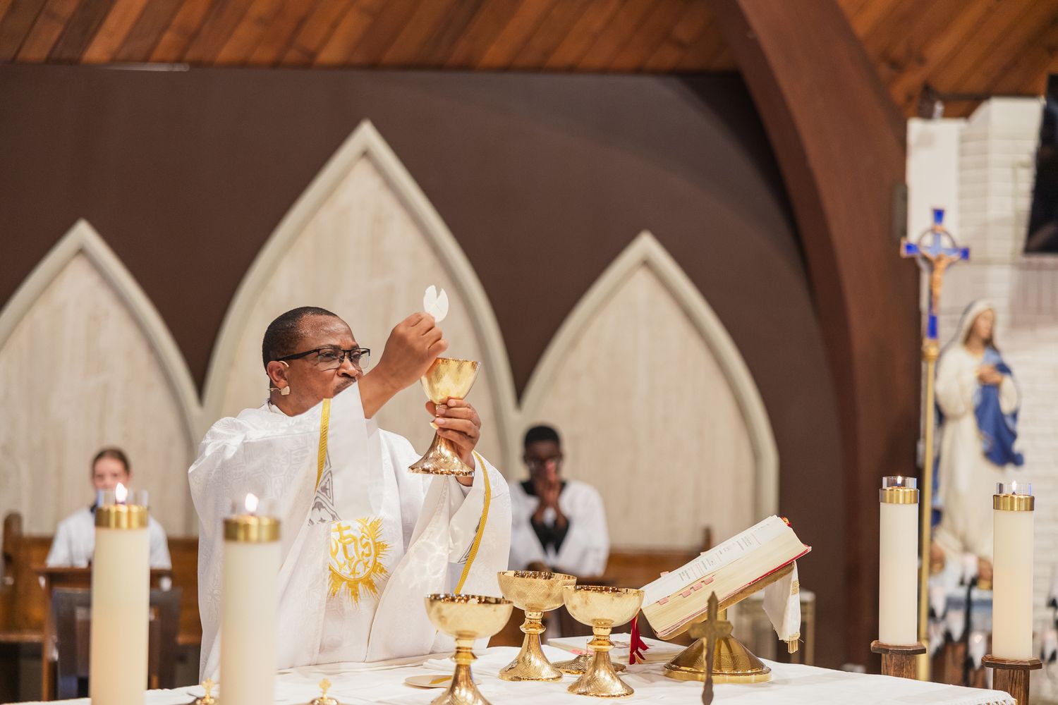Catholic priest elevates the Eucharist during Holy Mass at an ornate church altar with golden chalices.