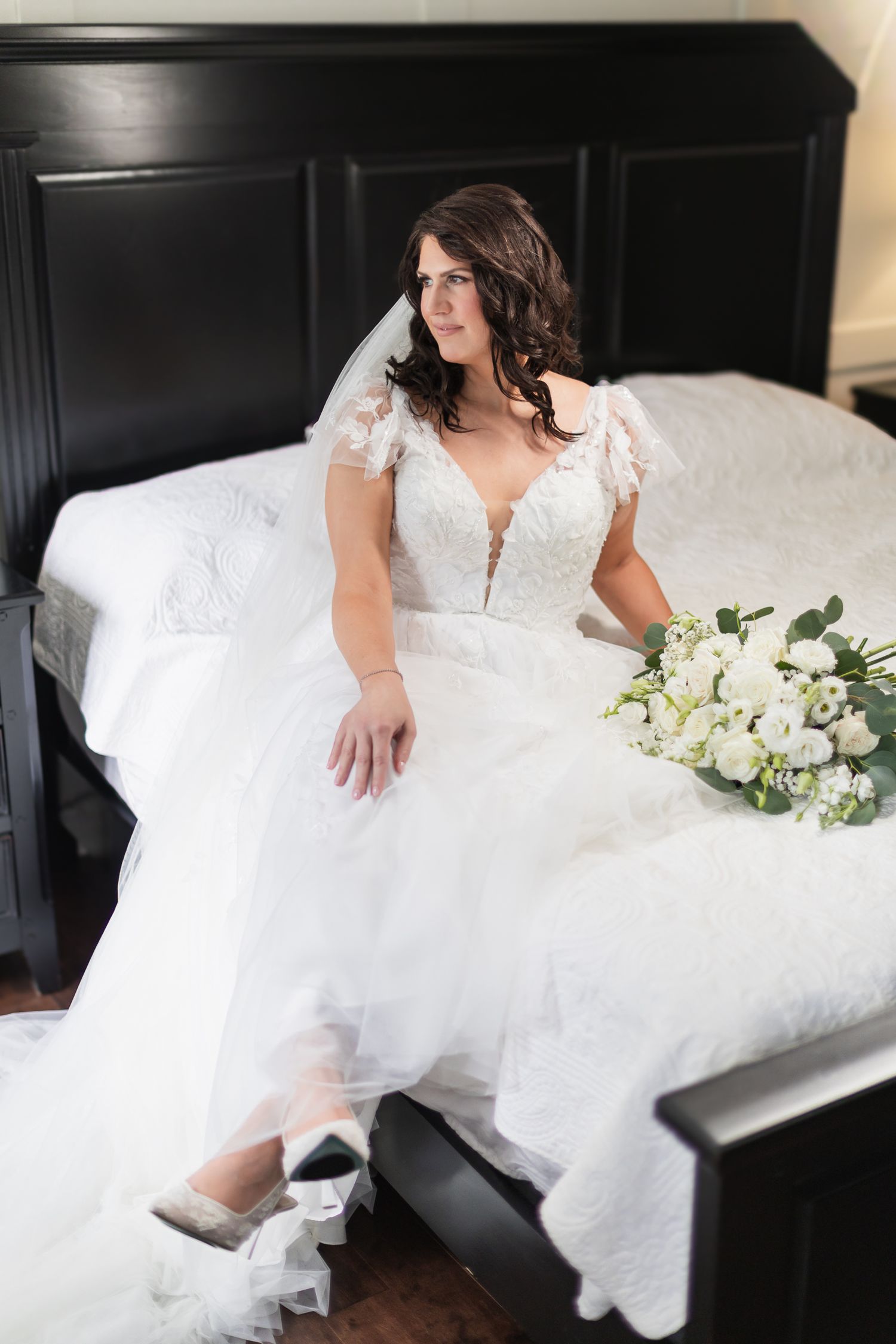 Bride in white lace ball gown sitting on bed with white floral bouquet, wearing veil and silver heels.