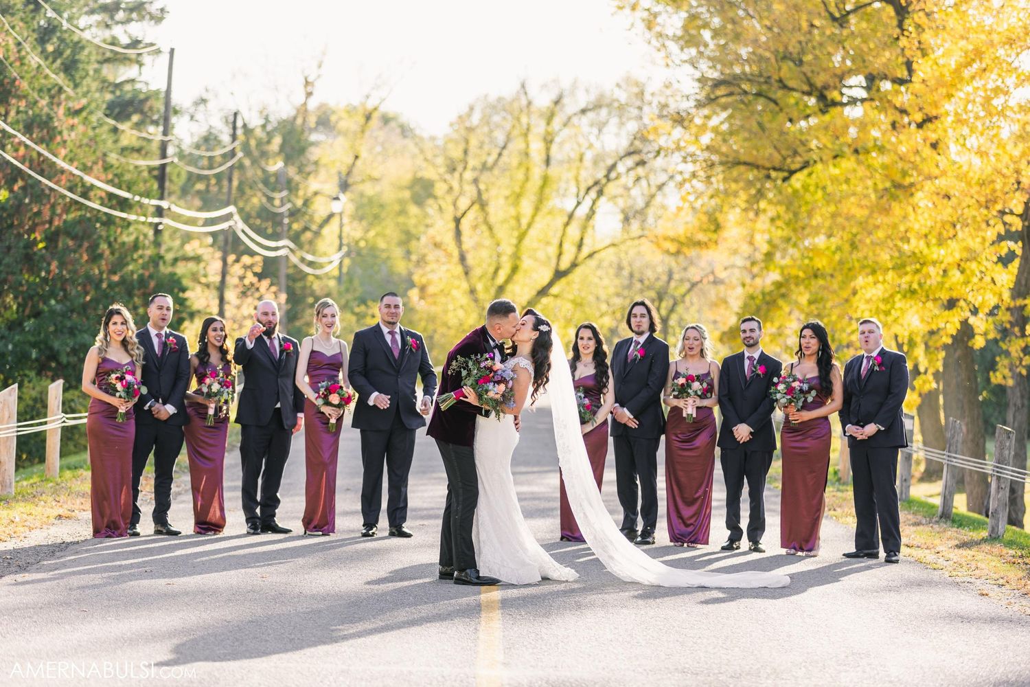 Wedding party in burgundy attire poses on tree-lined road during autumn with yellow foliage in background.