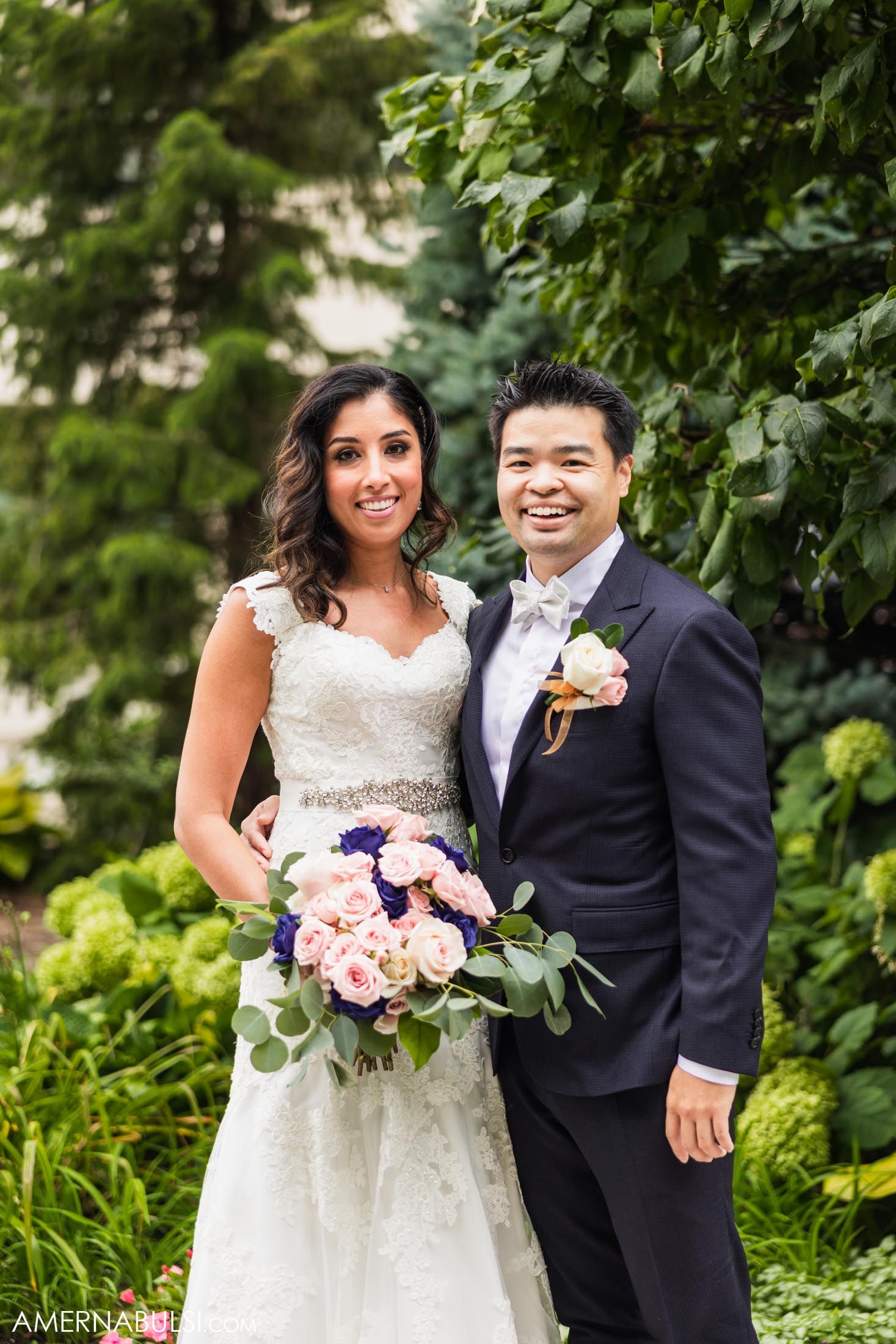 Newlyweds pose in lush garden setting with pink and purple bouquet.