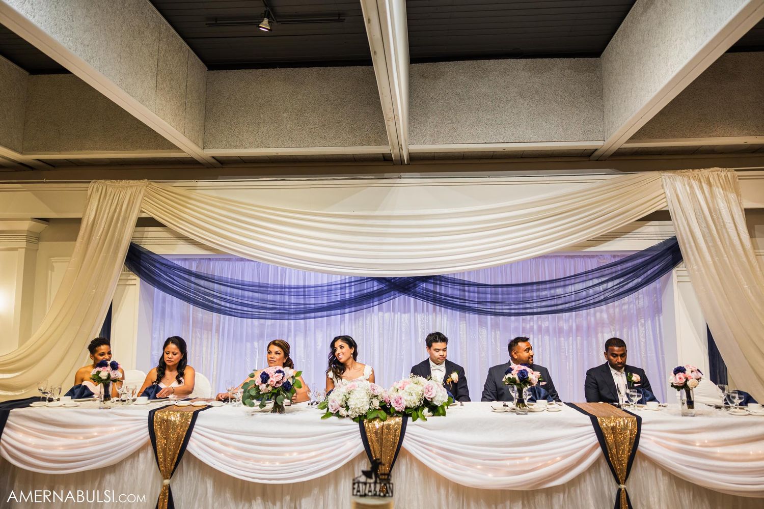 Wedding party seated at an elegant head table with white and blue drapery at a formal reception.