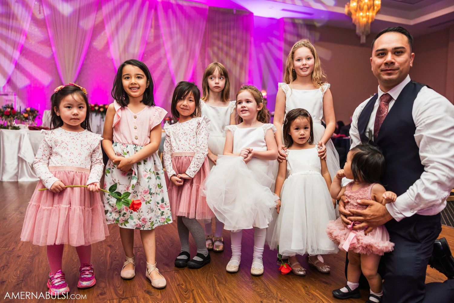 Young flower girls in pink and white dresses lined up with adults at wedding reception.