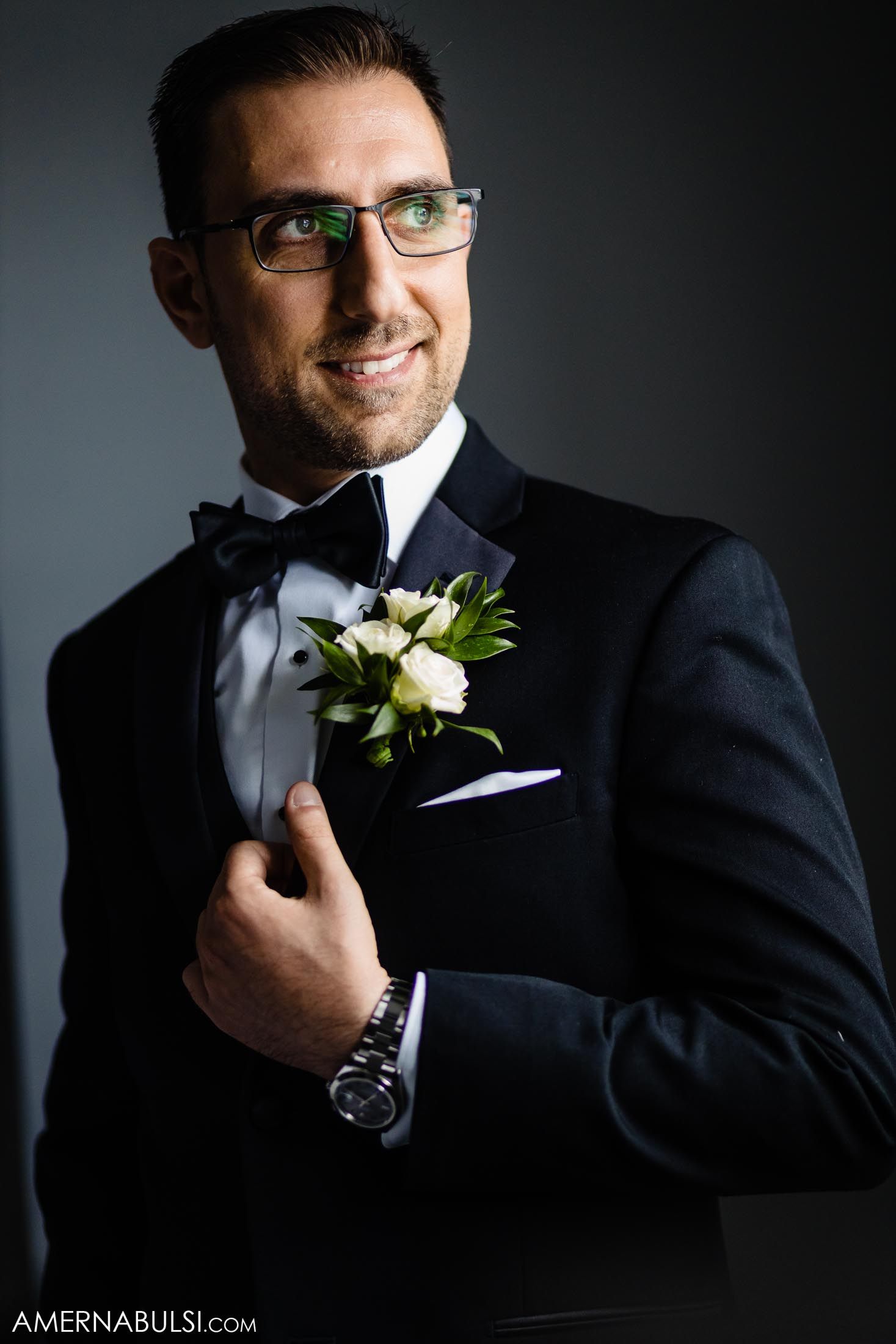 Groom wearing black tuxedo and glasses with white boutonniere smiling in dramatic lighting.
