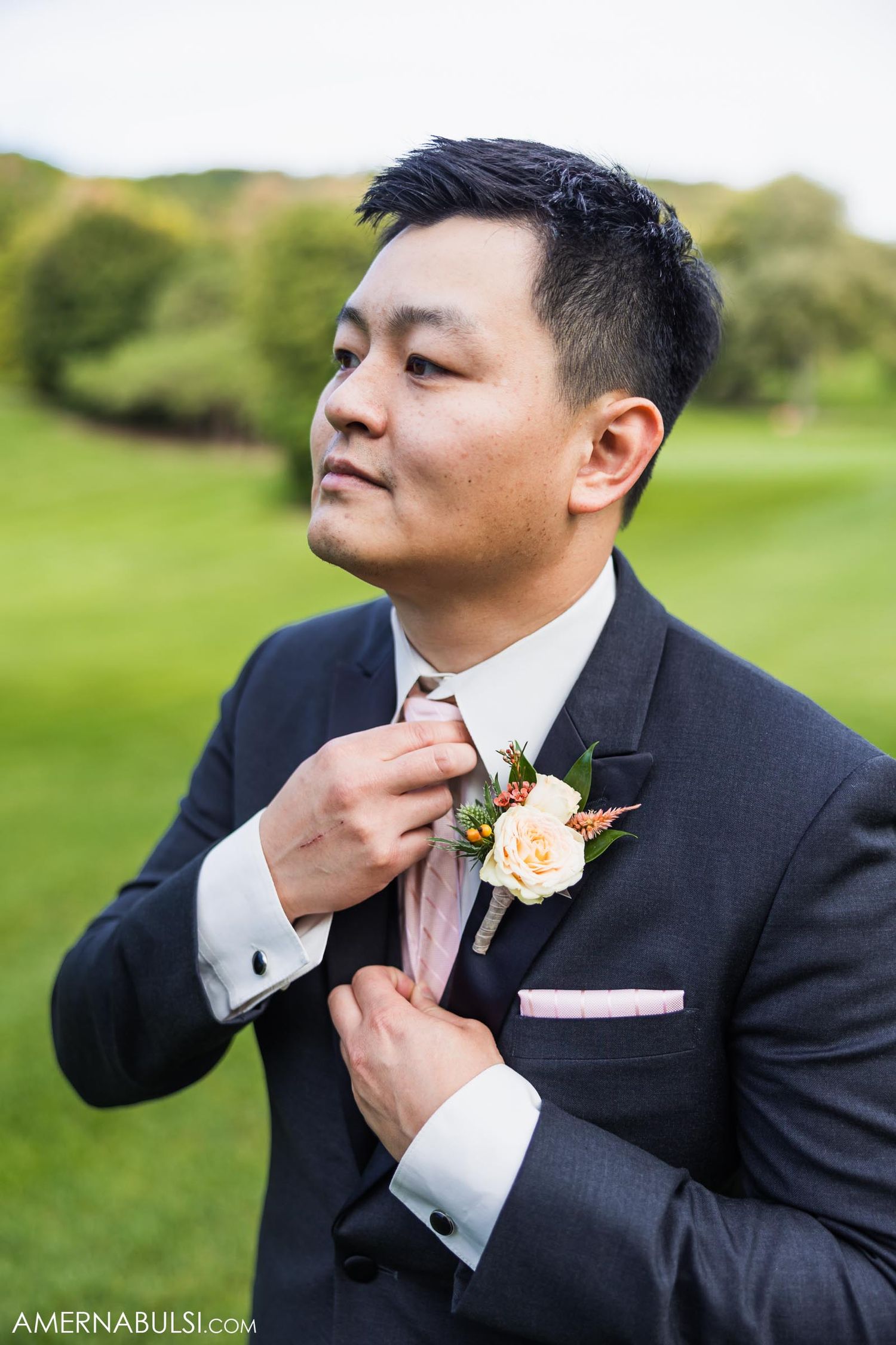 A groom in a dark suit adjusts his pink tie and boutonniere in an outdoor garden setting.
