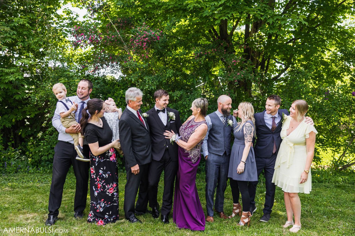 Large wedding party gathered outdoors for group photos among greenery.