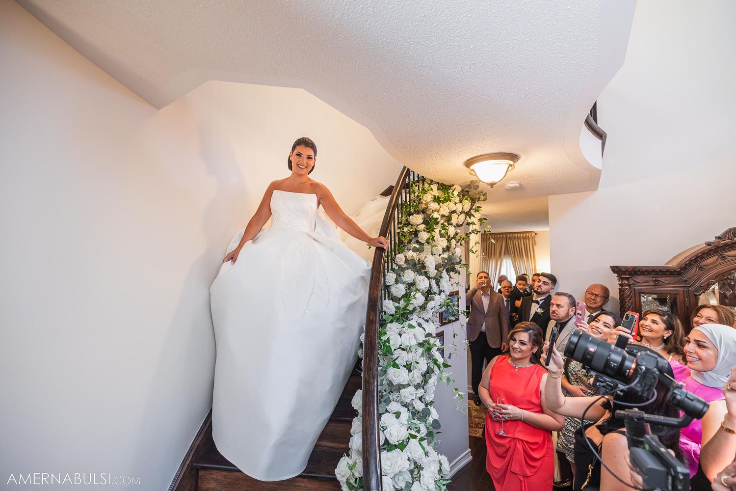 Bride in flowing white gown makes dramatic entrance down staircase to awaiting guests.