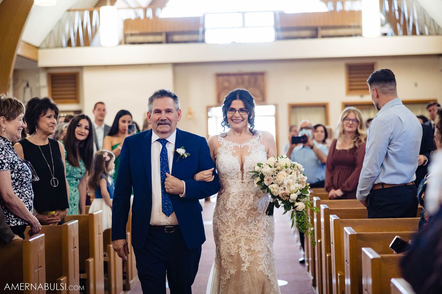 Father walks bride in champagne lace gown down church aisle as wedding guests look on.