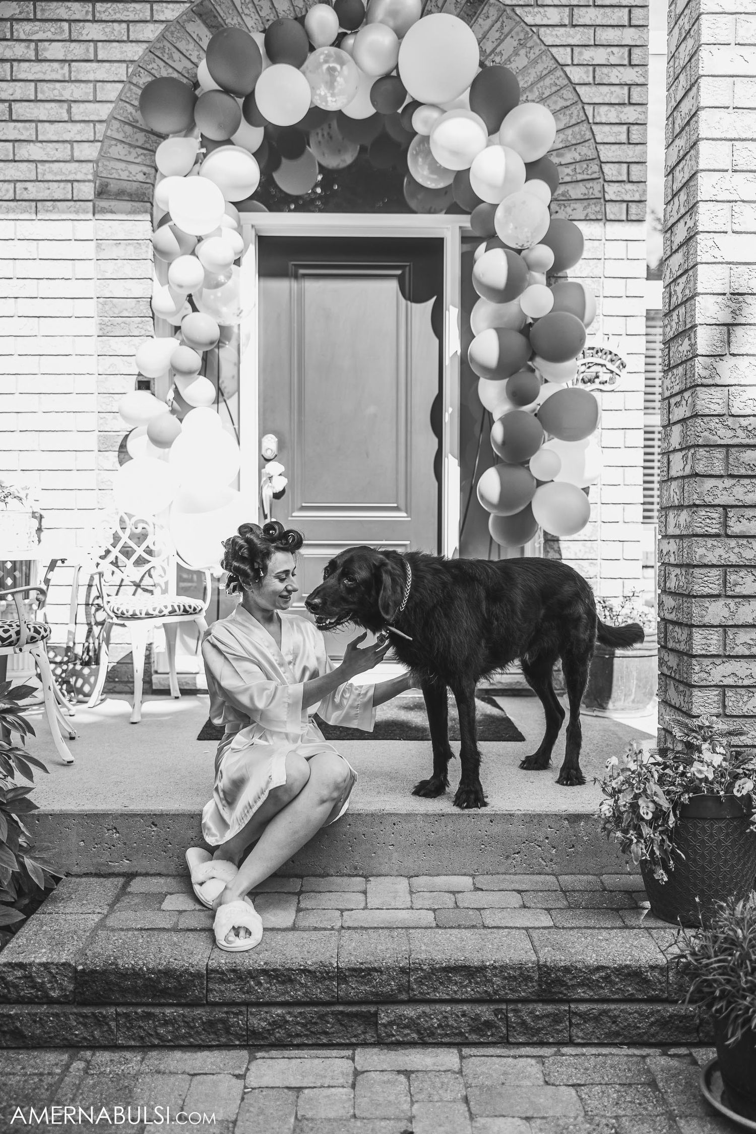 Black and white photo showing a person in white sitting with a large black dog under a balloon arch entryway.