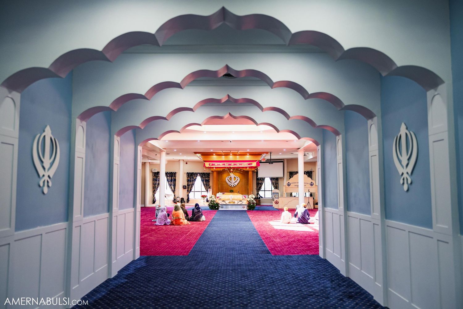 Interior view of a Sikh temple corridor with blue arches and red carpet leading to the prayer hall.