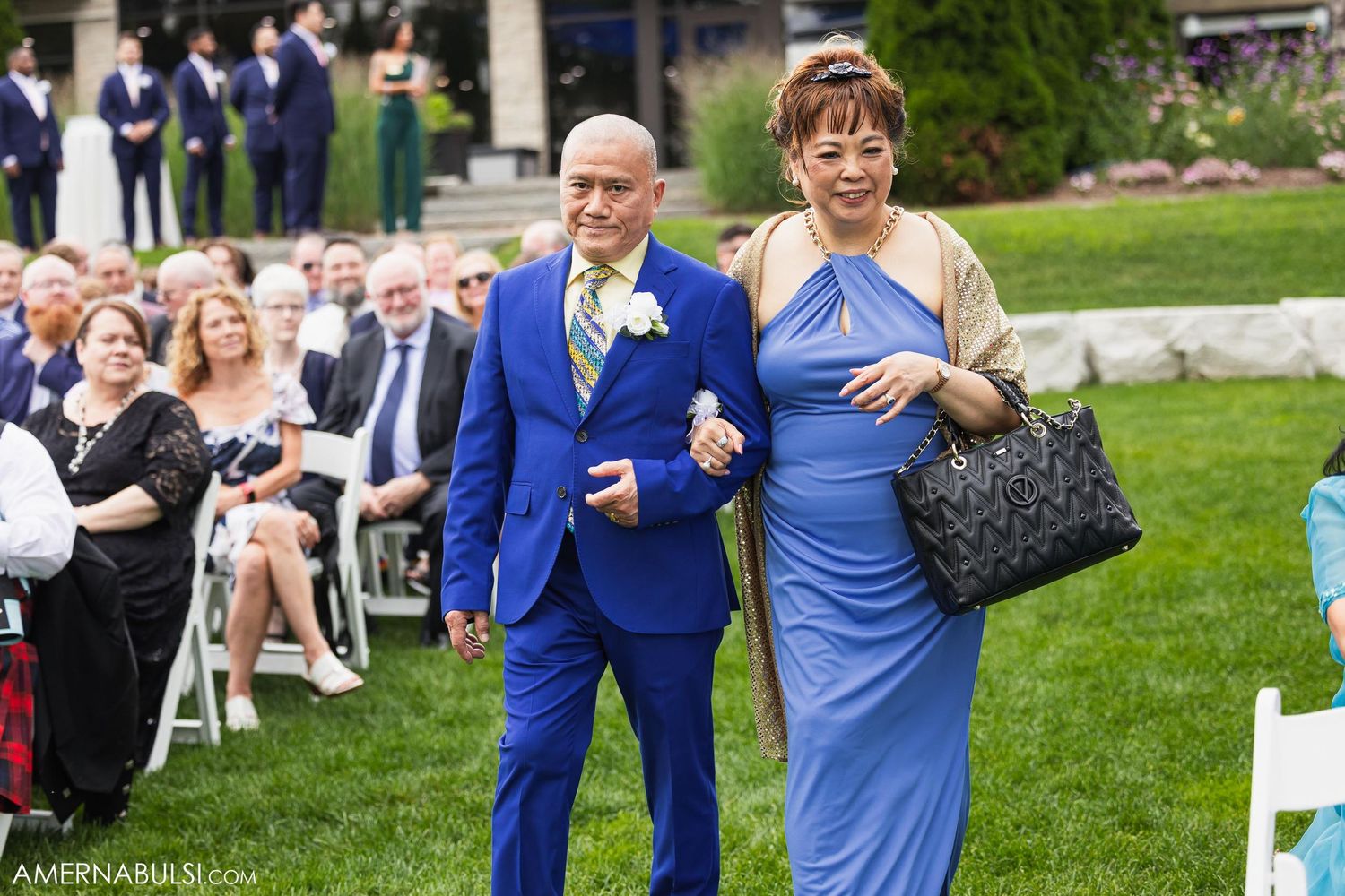 Wedding guests walk down aisle at outdoor ceremony with crowd seated on lawn.
