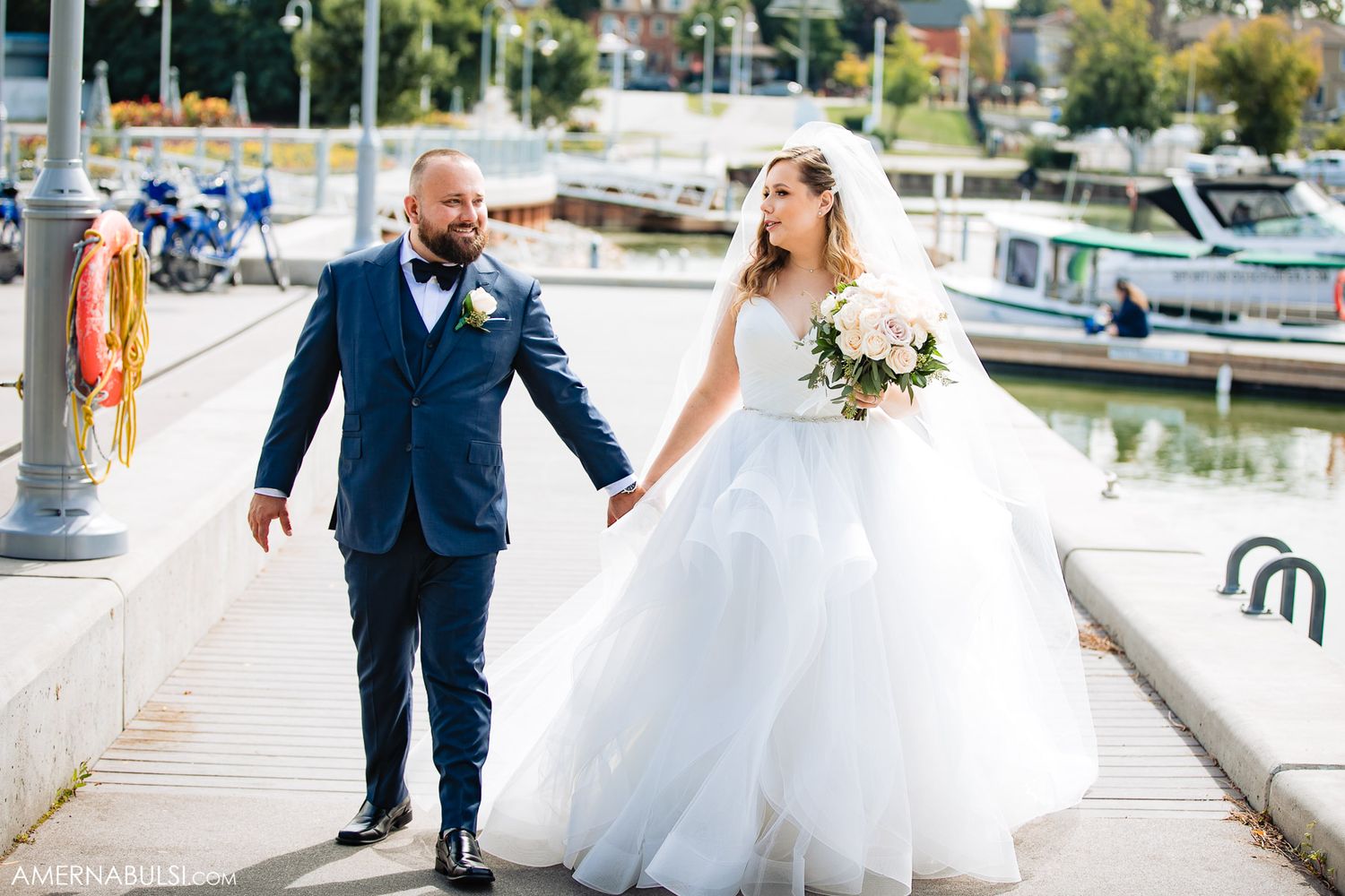 Wedding couple walks along a marina boardwalk with boats and water in the background.