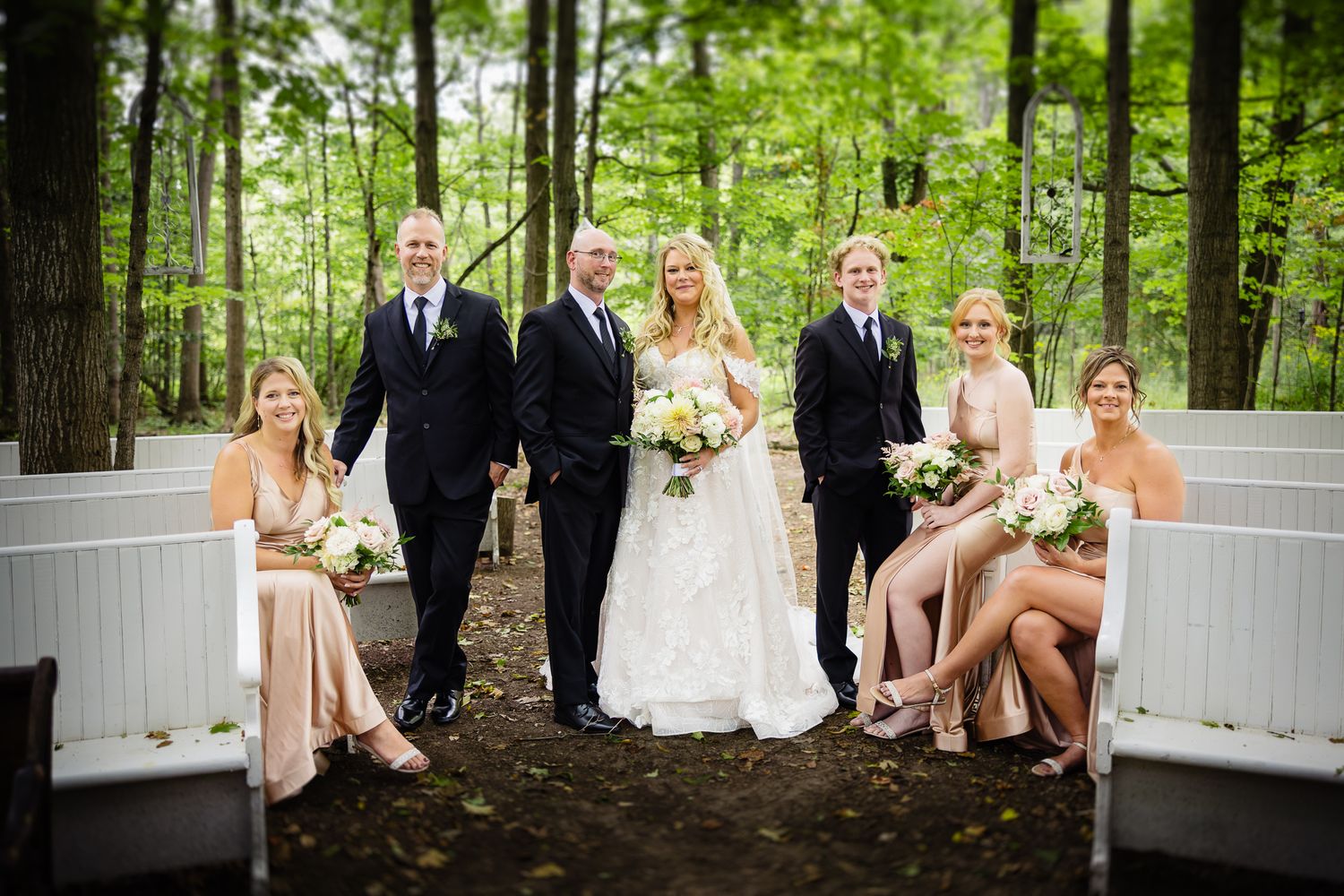 Wedding party poses on white benches surrounded by lush green forest setting during outdoor photo session.
