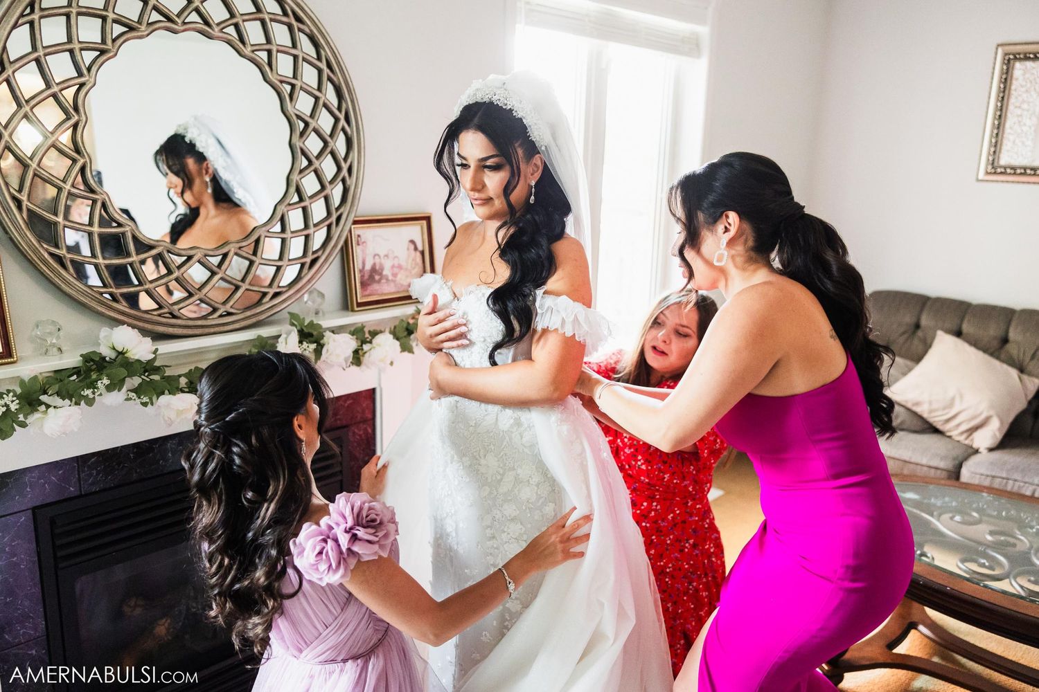 Bridesmaids in colorful dresses assist bride with final wedding preparations in front of circular mirror.
