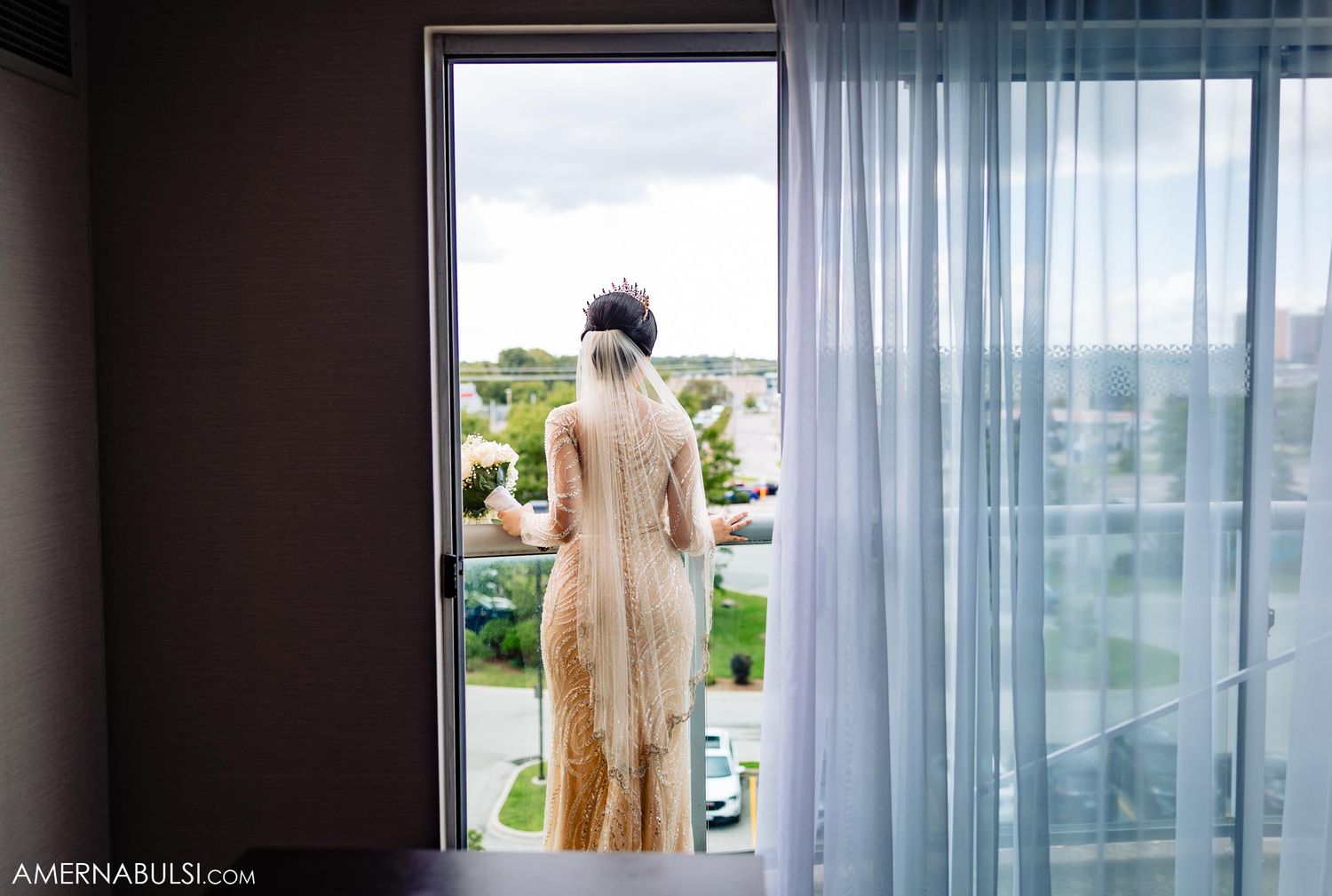 Silhouette of bride looking through window with flowing curtains and outdoor view.