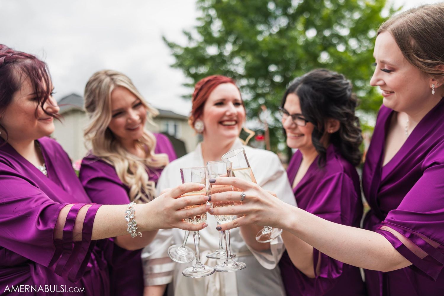 Wedding party in purple robes toasting with champagne glasses outside.