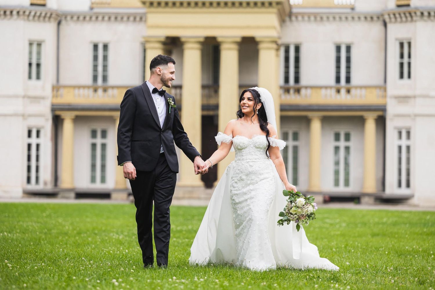 Newlyweds walking together on grass in front of a grand mansion estate.