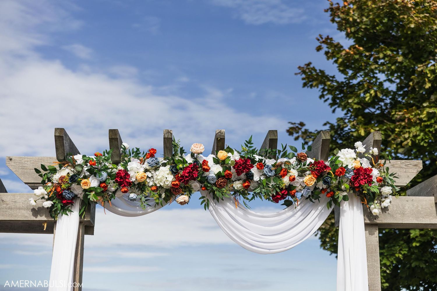 Wedding arch decorated with red and white flowers and white fabric against blue sky.