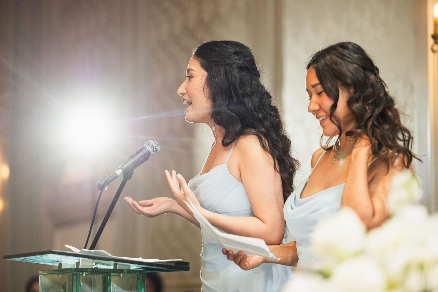 Bridesmaids in light blue dresses giving speeches at wedding reception podium.