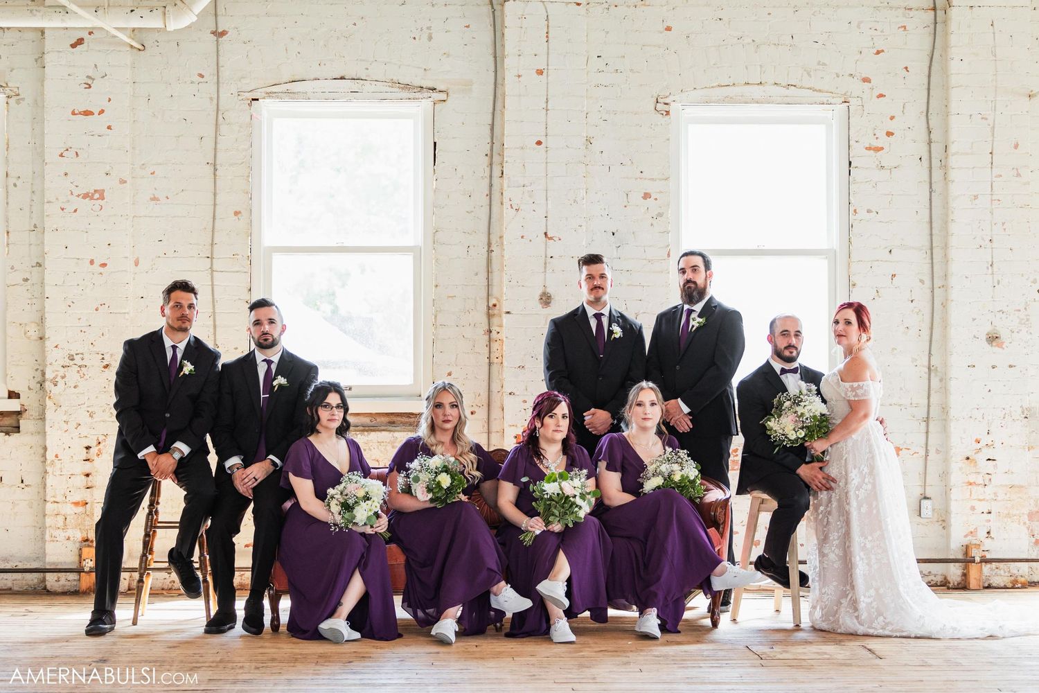 Wedding party in purple and black attire posed together in bright industrial space with brick walls.