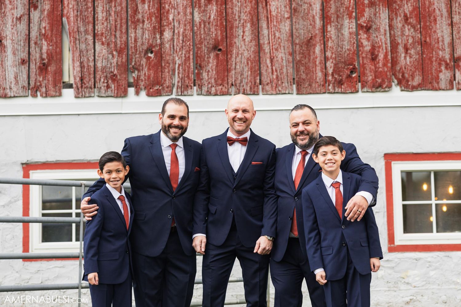 Groomsmen and young boys pose for formal photo in matching navy suits against rustic red barn.