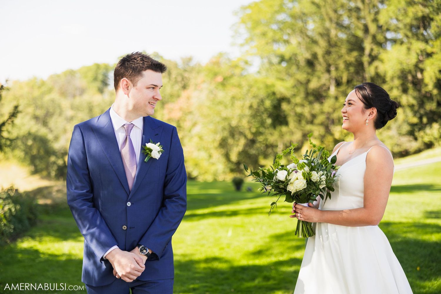 Wedding couple stands together on sunny lawn with bride holding white and green bouquet.