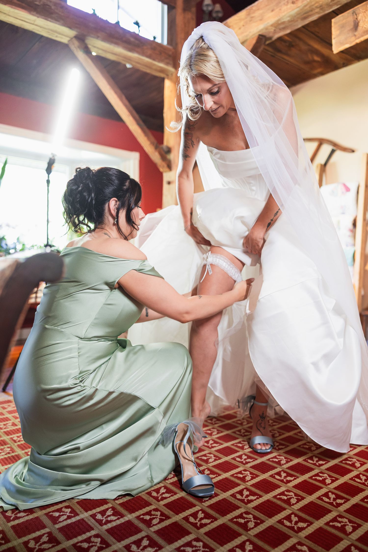 Bridesmaid in sage green gown helps bride put on garter before wedding ceremony indoors.