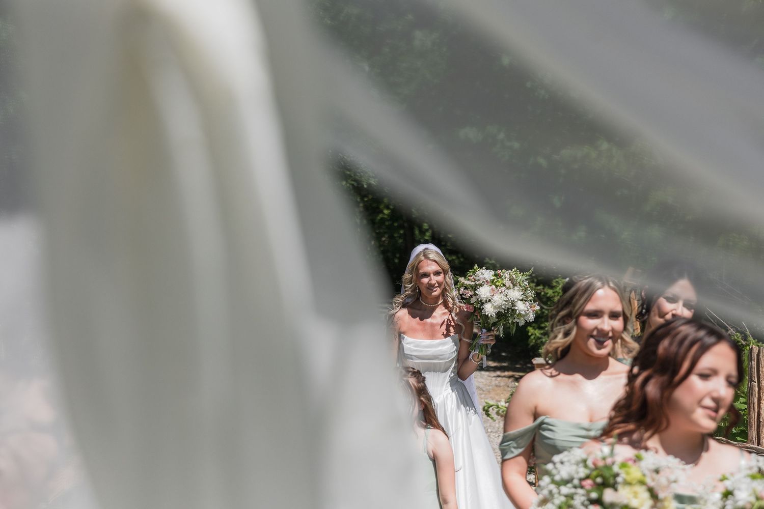 Bride in white gown walks with bridesmaids in sage green dresses, captured through flowing veil outdoors.