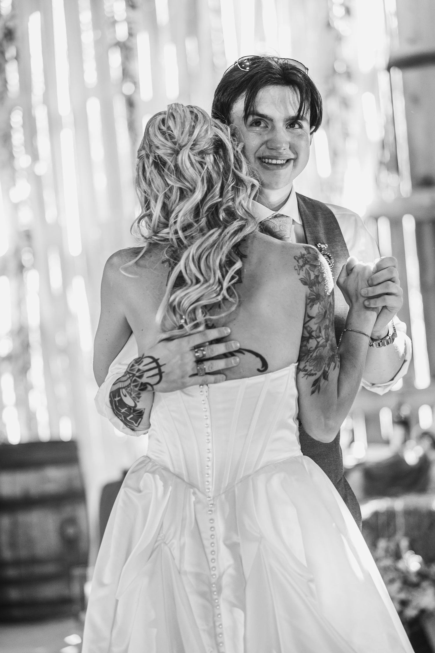Black and white photo of a groom with tattoos smiling while sharing first dance with bride in white gown at wedding.