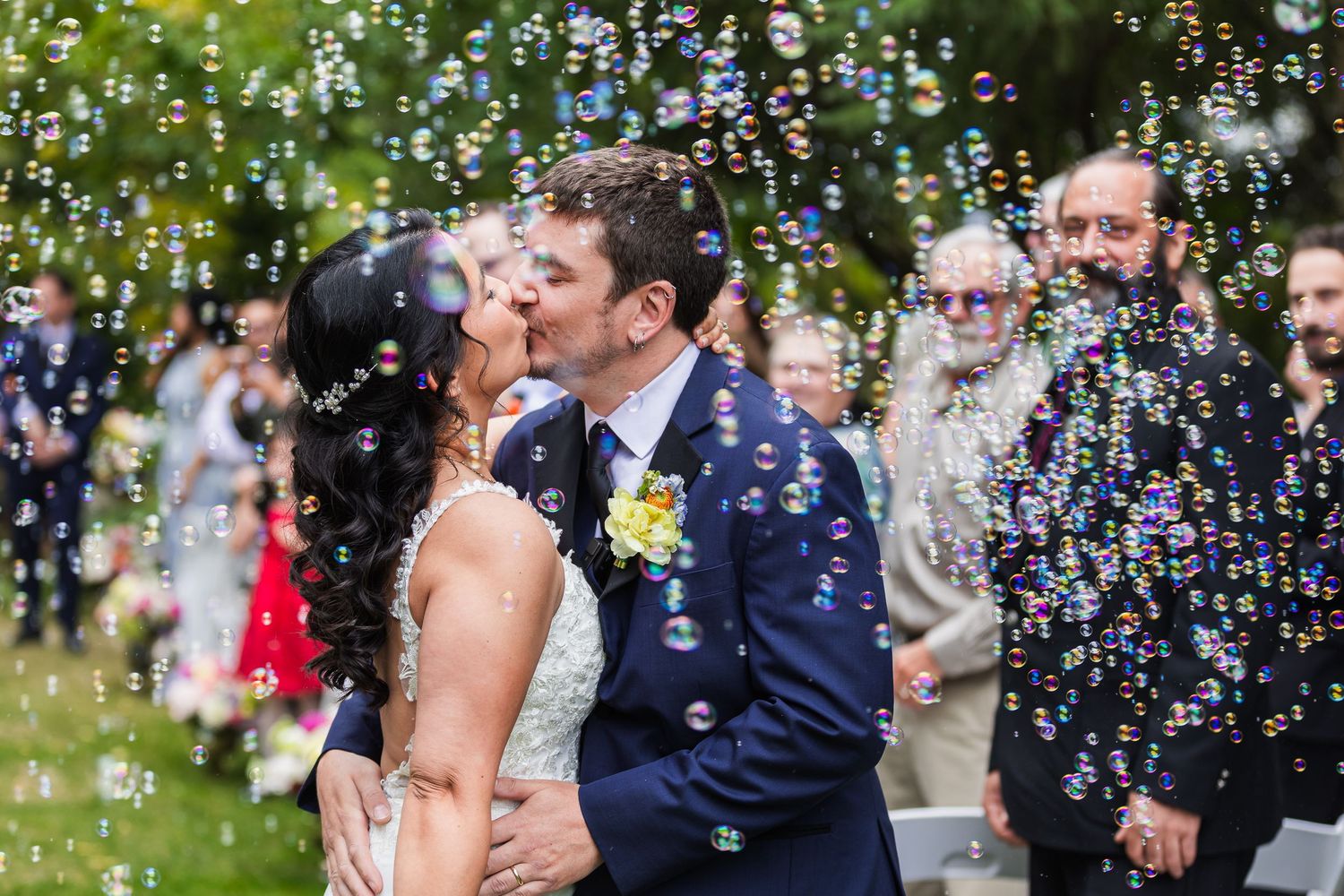 Newlyweds share a first kiss surrounded by bubbles at an outdoor wedding ceremony with guests watching.