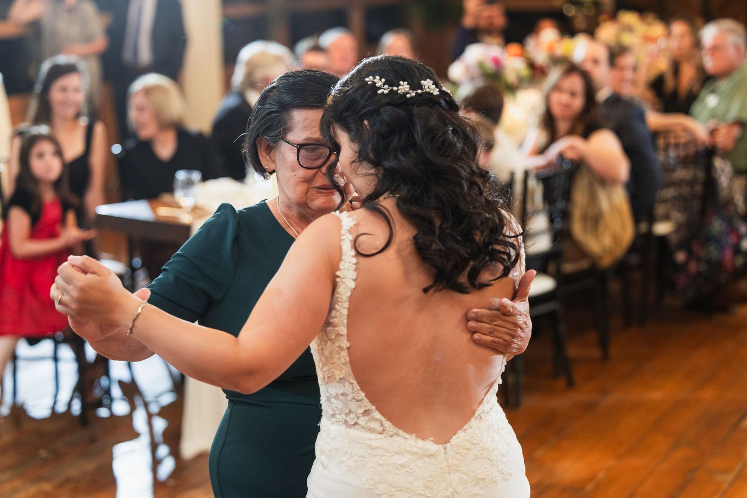 Bride sharing an emotional first dance with a family member in a lace wedding gown at a rustic reception venue.