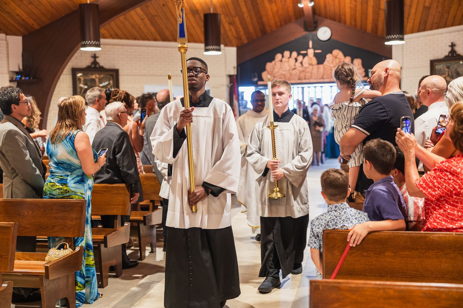 Clergy processing down the aisle during a Catholic church ceremony with congregation standing in wooden pews.