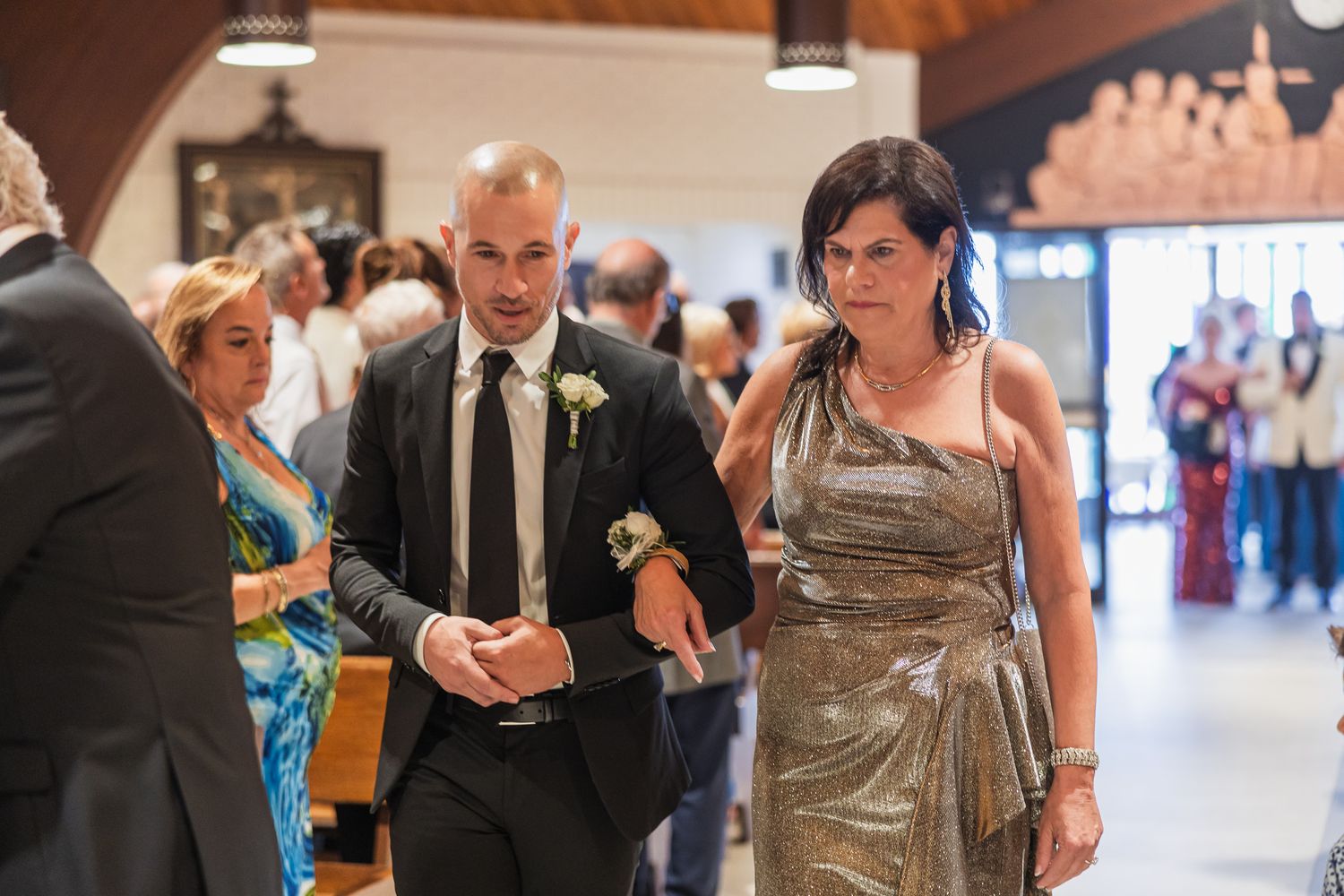 Groom escorted by woman in gold dress walking down church aisle during wedding ceremony.