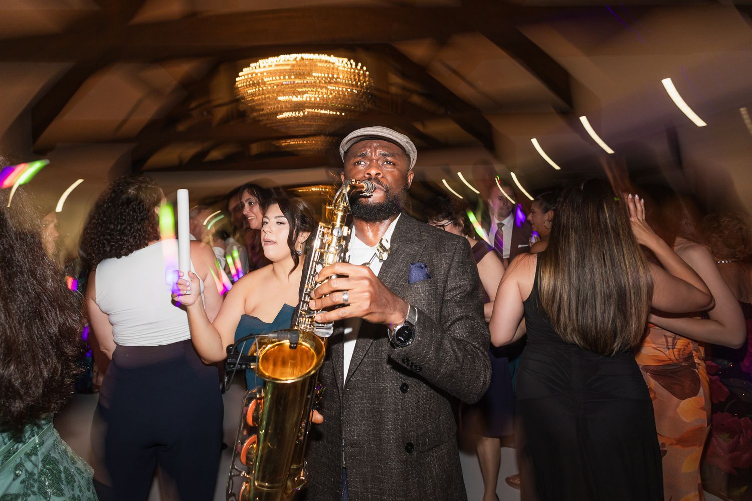 Saxophonist performing at a lively wedding reception party with guests dancing under warm chandelier lighting.